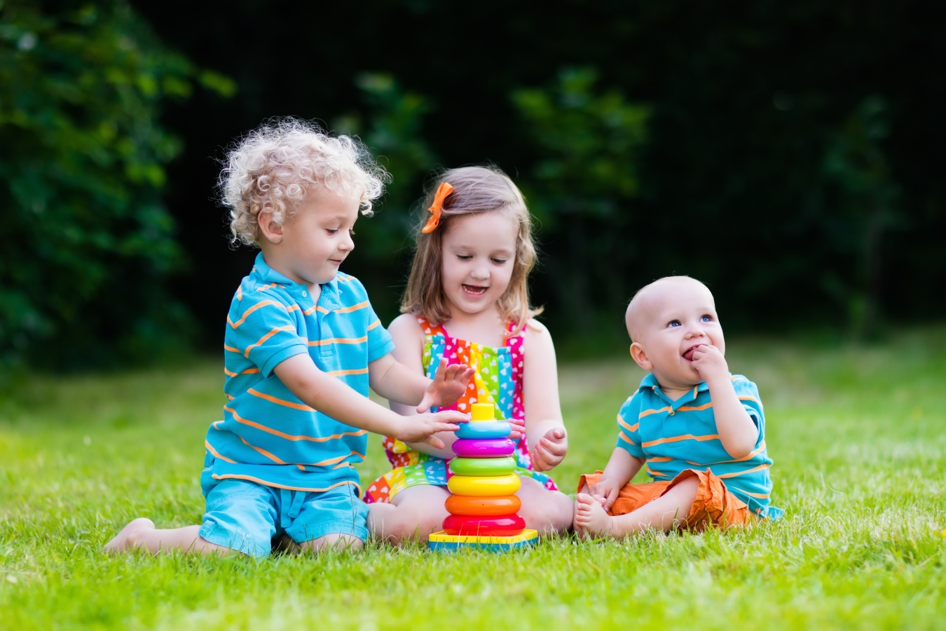 A brother and sister and their baby brother play stacking rings together on the grass