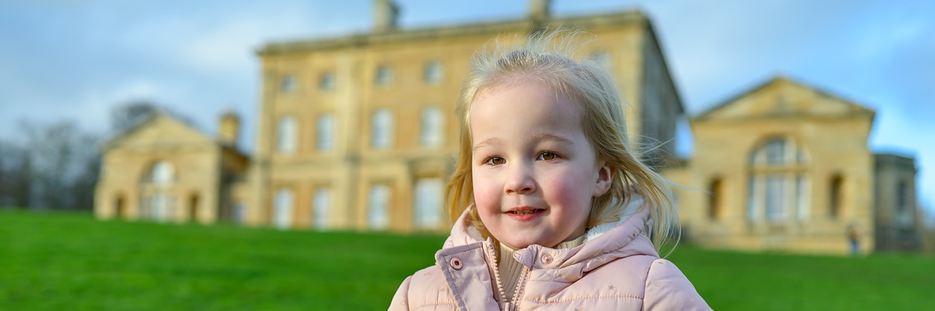 A little girl with blonde hair and pink coat holding a teddy in front of Cusworth Hall