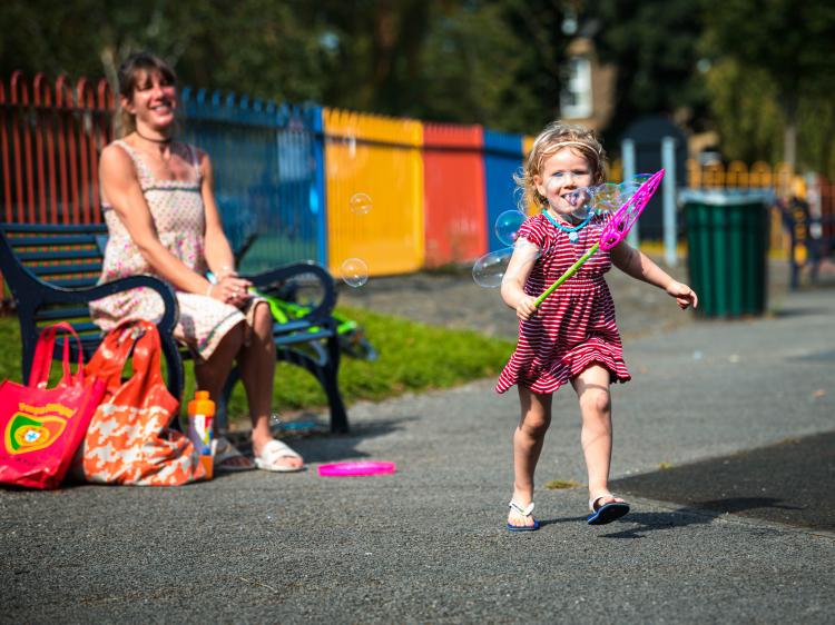 Happy mum on bench watching young daughter running with bubble stick