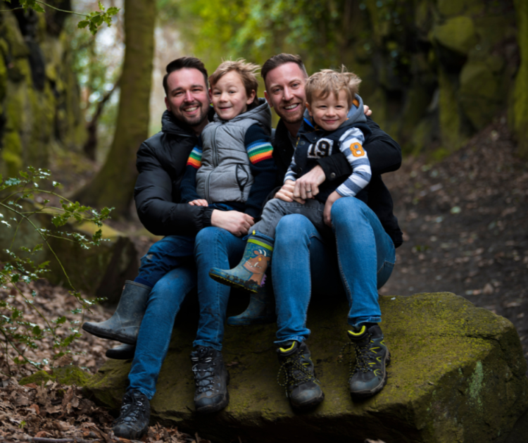 two dads with two little boys sitting on their knees out for a walk in the woods
