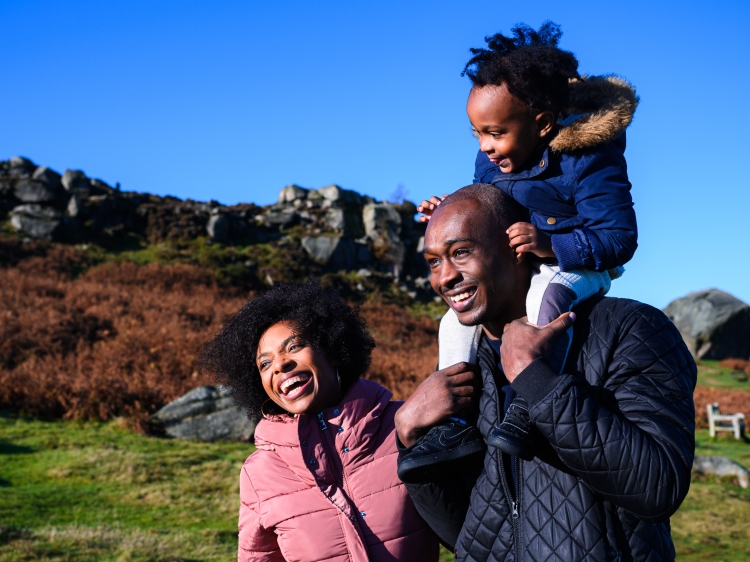 A laughing family with toddler on dad's shoulders walking across Ilkley Moor
