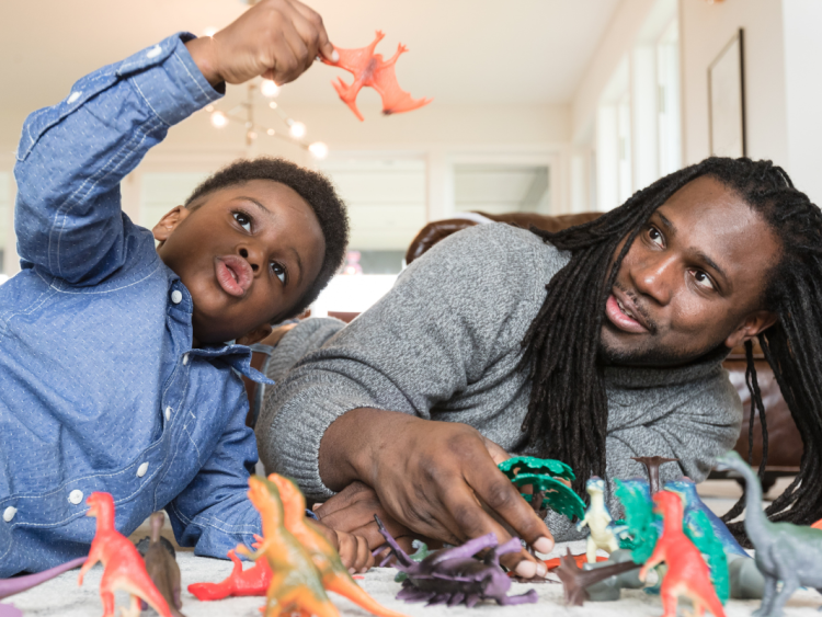 Dad with long dreadlocks and son lay on the floor playing with dinosaurs