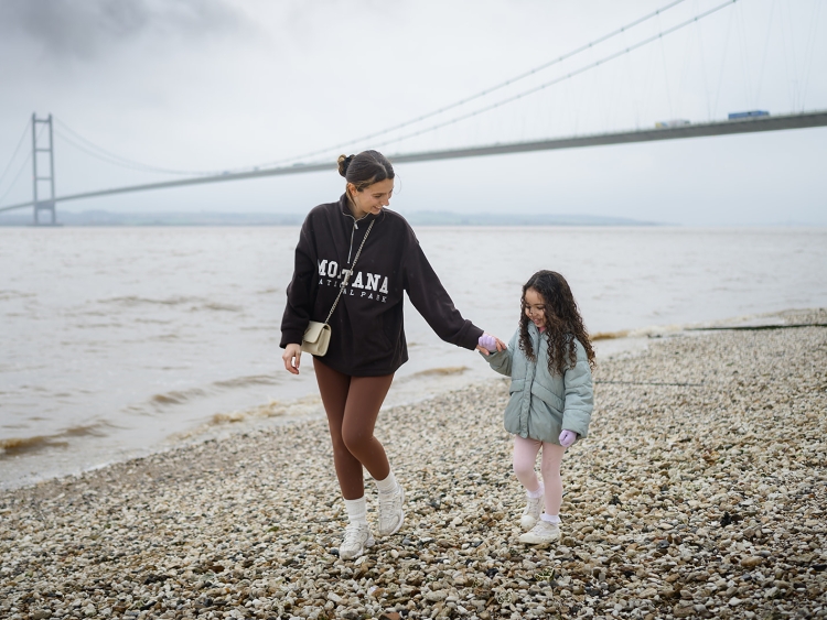A mother and daughter walking along a shingle beach with humber bridge in the background on a misty day