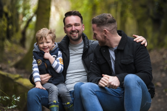 happy male couple with young son on knee in woods