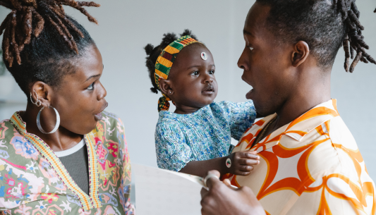 Mum and dad both with dreadlocks pulling excited faces at their baby girl who has a sticker on her head in a playful way