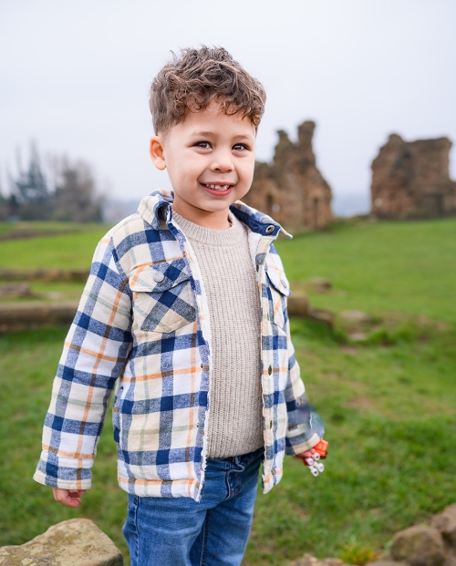 Little boy with curly hair and checked jacket outside with sandal castle in background